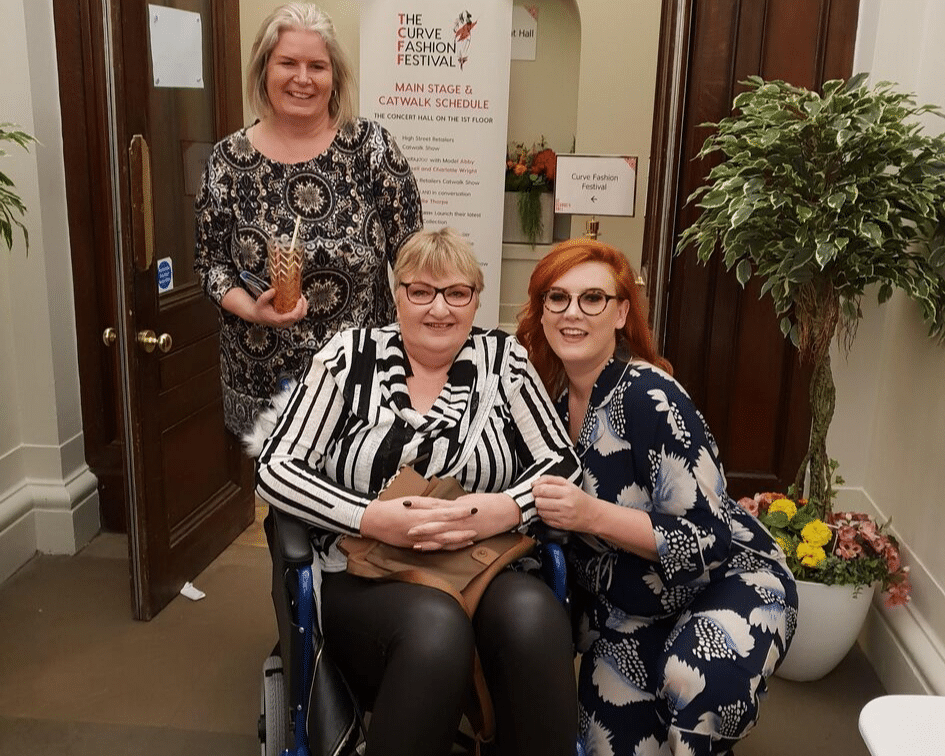 Rachael in a black and white jumper with black leggings, with Emma at her side and Jackie stood behind her in a transit wheelchair